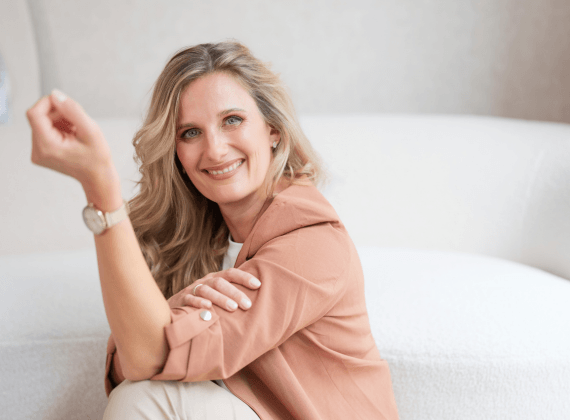 Doreen von La Webista sitzt lächelnd vor einem hellen Sofa und blickt offen in die Kamera